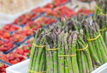 Bottes d'asperges vertes fraîches sur un étal au Marché Pernoud, avec des barquettes de fraises en arrière-plan, pour un mois de mars savoureux.