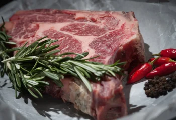 Pièce de viande crue avec du romarin, des piments rouges et des grains de poivre noir.