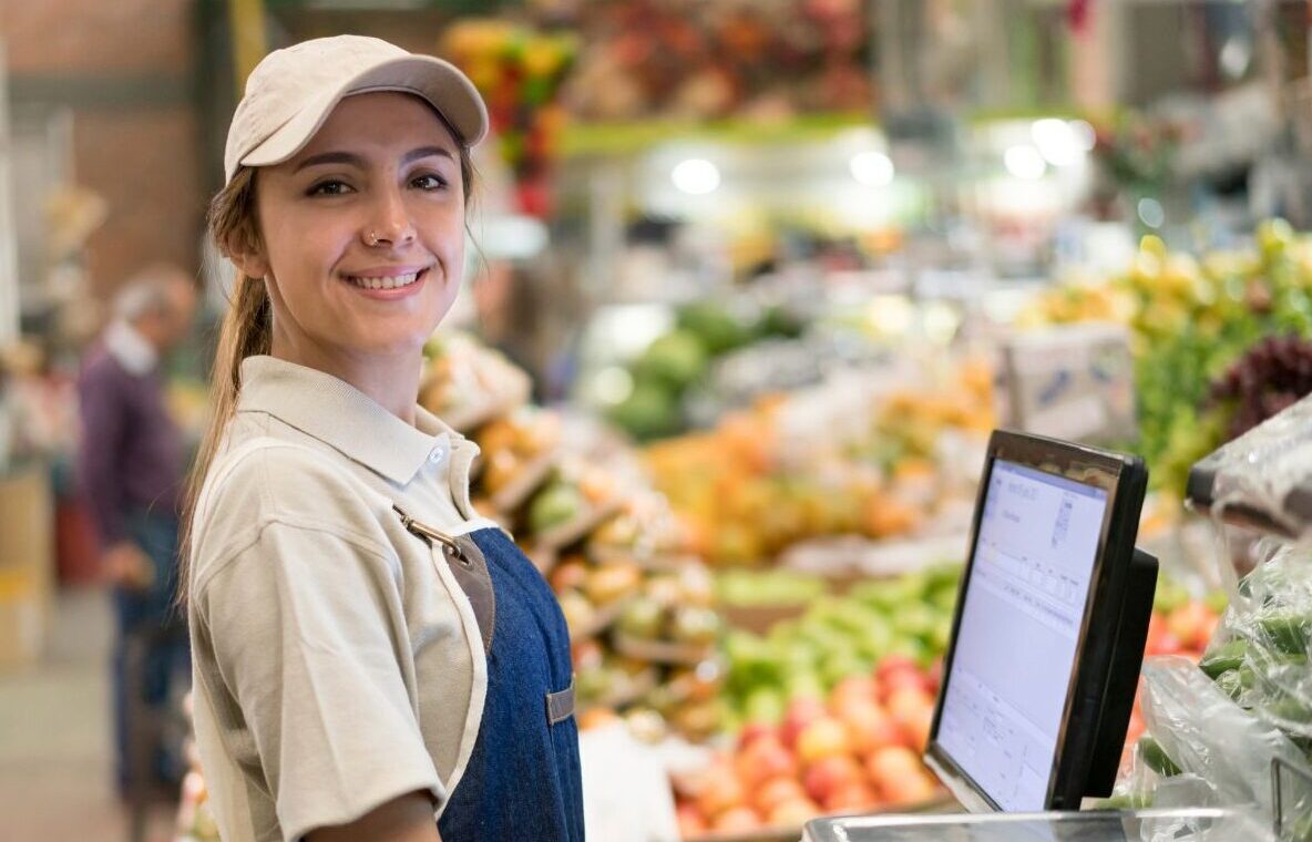 Caissière souriante au Marché Pernoud, prête à servir les clients, devant un étalage de fruits et légumes frais.