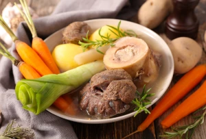 Plat de pot-au-feu traditionnel avec des morceaux de viande, des os à moelle, des carottes, des poireaux et des pommes de terre, joliment disposés dans une assiette blanche, accompagné de branches de romarin pour la décoration.