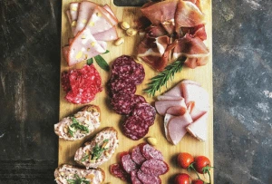 Planche de charcuterie sur une planche en bois, avec une variété de viandes tranchées comme le jambon cru, la mortadelle, le salami et d'autres saucissons, accompagnée de quelques pistaches, tomates cerises et branches de romarin.